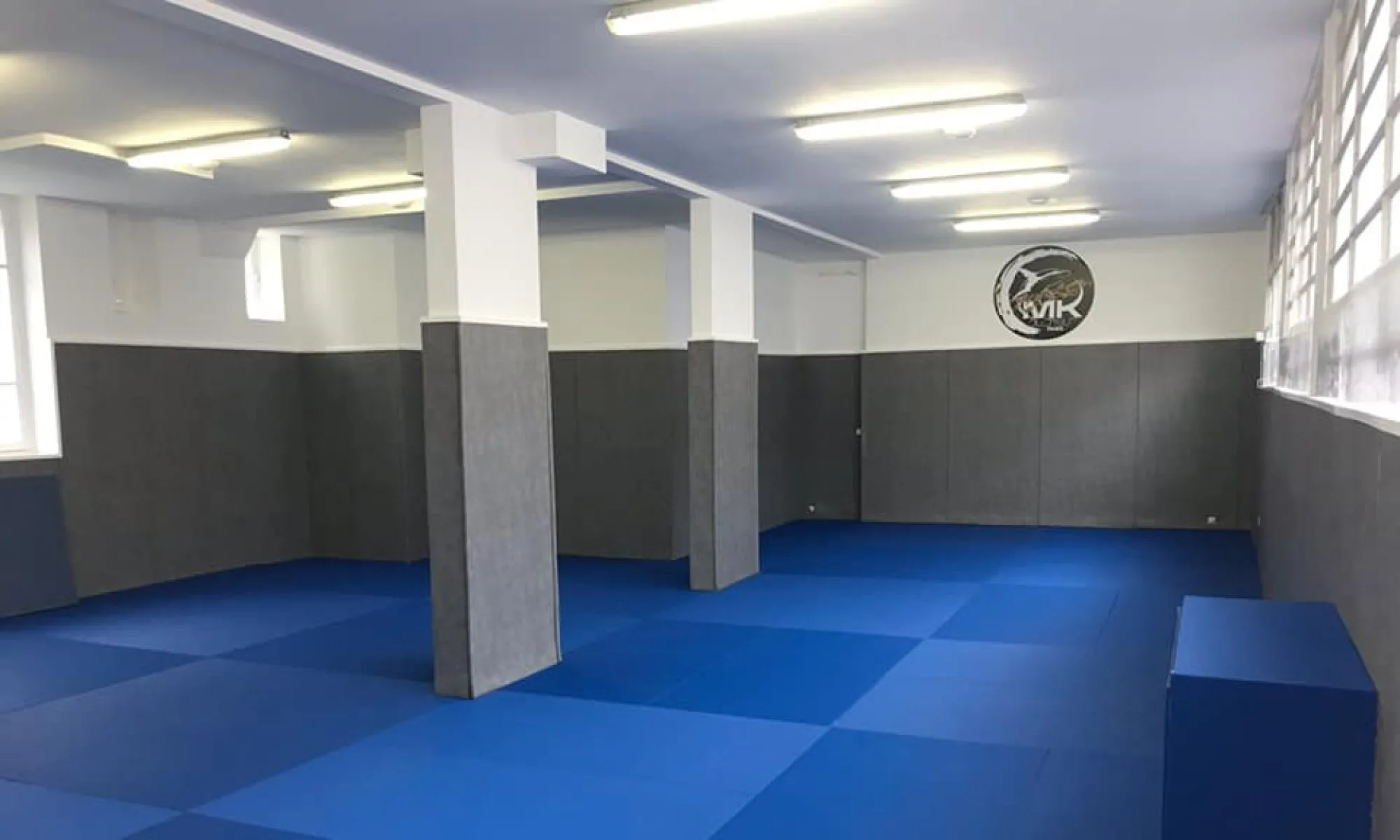 Le dojo vide : tatamis bleus, murs capitonnés et logo MK au mur